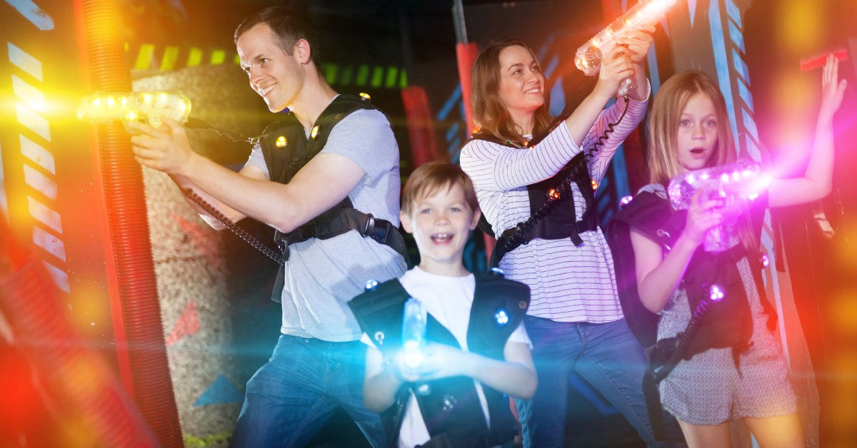 Two adults play laser tag with two kids in a dark room with neon accents. The players' laser guns and vests glow colorfully.