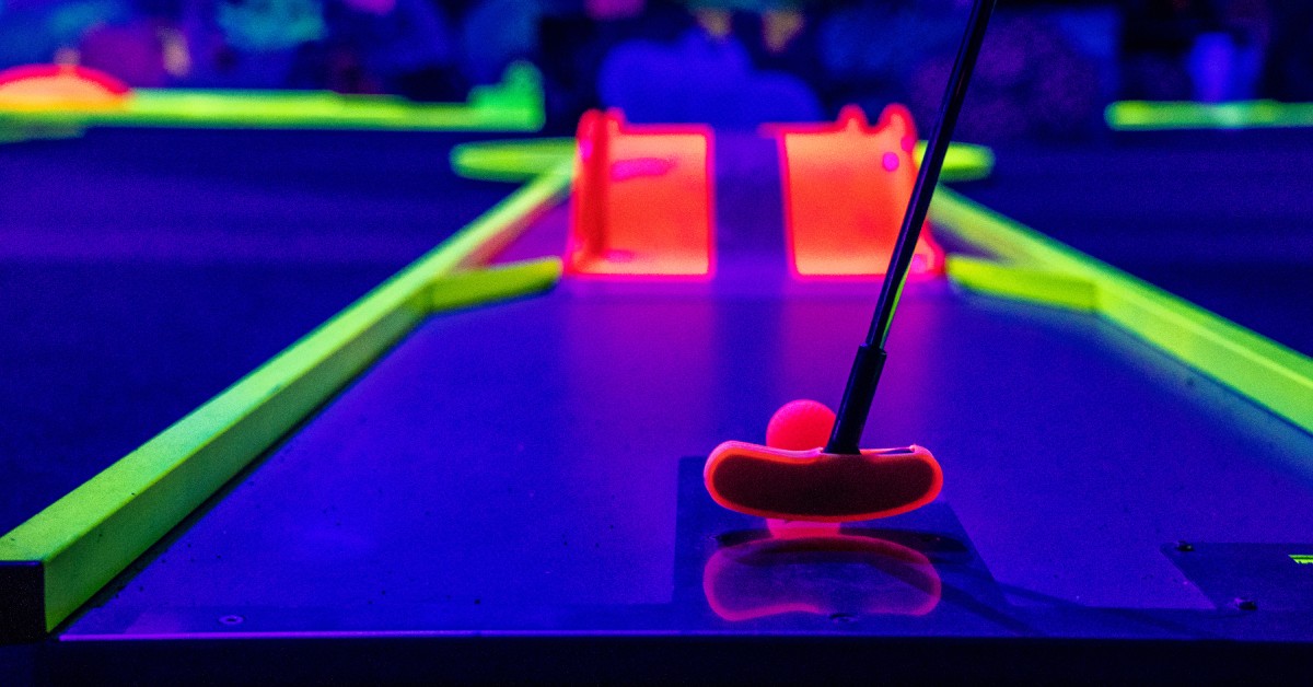 A close-up of a mini golf putter positioned to hit a ball over a ramp. The course is lit by black light and glows in neon colors.