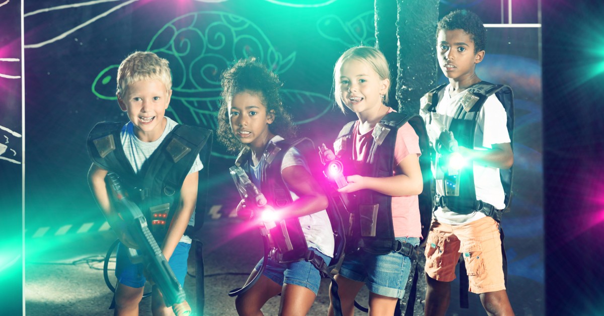 Four kids standing together in a dark laser tag arena. They each wear a vest and hold a laser gun that emits a bright light.