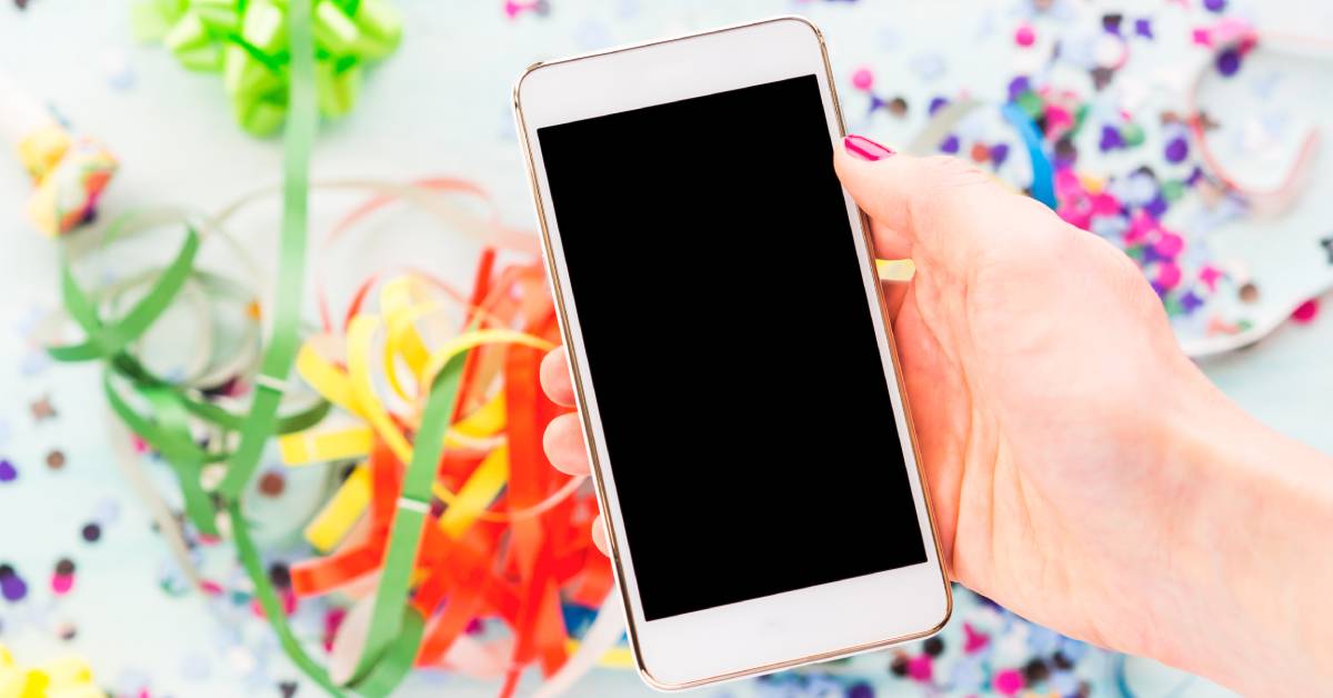 A person holds a cell phone above party streamers, confetti, and gift bows. The cell phone screen is black.