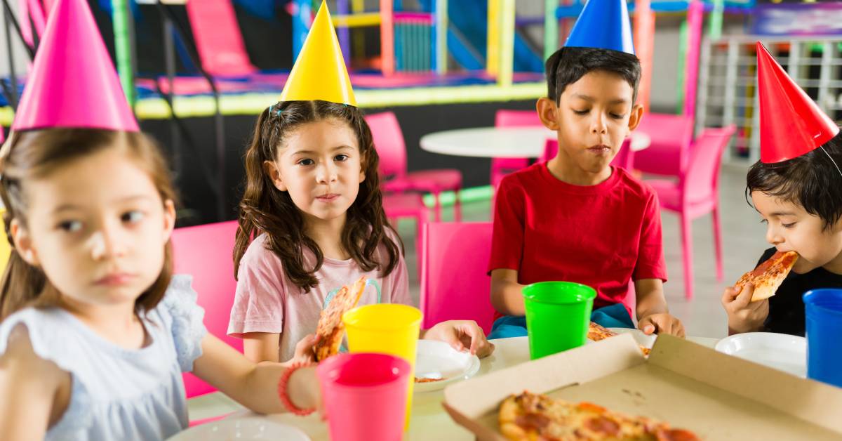 Kids wearing party hats sit at a table and eat pepperoni pizza. Behind the kids is a colorful play place and another table.