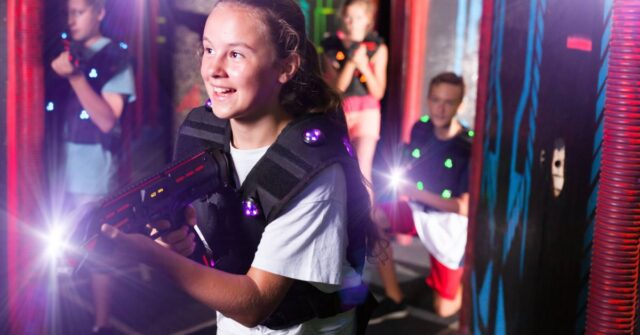 A smiling child holds her laser tag gun in both hands as she moves to her right. Three other child-aged players wait behind her.