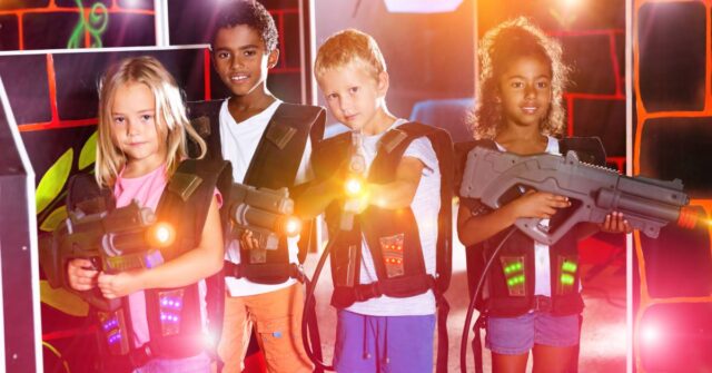 Four young kids stand together in a room lit with neon lights. They hold their laser tag guns pointed in different directions.